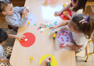 Dzieci siedzą przy stoliku i wykonują prace plastyczną z użyciem kolorowych kropek.