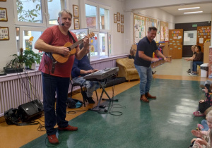 Zdjęcie przedstawia dwóch Panow. Jeden gra na ukulele, a drugi kleszcze w rytm muzyki