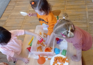 Zdjęcie przedstawia trzy dziewczynki podczas zabawy sensorycznej z marchewką i wodą.