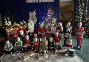 Cała grupa pozuje z Mikołajem i prezentami na tle dekoracji i oświetlonych choinek