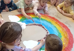 Zdjęcie przedstawia dzieci malujące farbami kolor fioletowy na tęczy.