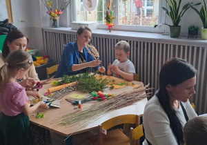 Dzieci z rodzicami siedzą przy stolikach i tworzą palmę Wielkanocną.