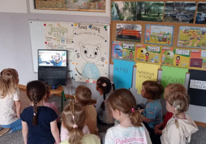 Zdjęcie przedstawia dzieci siedzące na dywanie, zgromadzone wokół laptopa. Słuchają czytanej bajki pt.: „Kicia Kocia. Co z tymi śmieciami?”.
