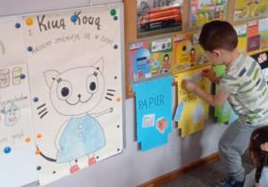 Zdjęcie przedstawia chłopca przyklejającego ilustrację gazety do niebieskiego kosza z napisem „papier”.