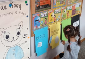 Zdjęcie przedstawia dziewczynkę przyklejającą ilustrację słomek do żółtego kosza z napisem „tworzywa sztuczne”.