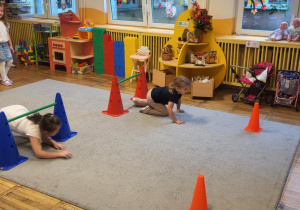 Dwie dziewczynki przechodzą pod przeszkodą ułożoną z pachołów i laski gimnastycznej.