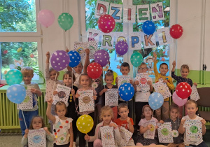 Dzieci pozują do zdjęcia grupowego z balonami w kropki i pracami plastycznymi.