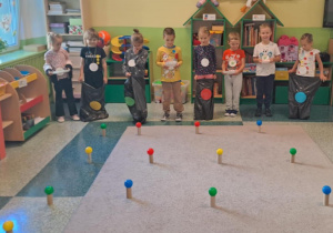 Dzieci stoją w rzędzie z założonymi workami w kolorowe kropki na dywanie rozłożone są kolorowe piłki na rolkach po papierze toaletowym.