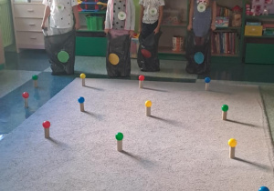 Dzieci stoją w rzędzie z założonymi workami w kolorowe kropki na dywanie rozłożone są kolorowe piłki na rolkach po papierze toaletowym.