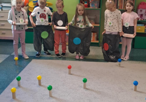 Dzieci stoją w rzędzie z założonymi workami w kolorowe kropki na dywanie rozłożone są kolorowe piłki na rolkach po papierze toaletowym.