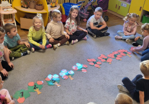 Dzieci siedzą na dywanie, dziewczynka liczy jabłka i dopasowuje do odpowiedniej cyfry na drzewie.