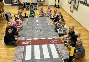 Dzieci siedzą przy macie edukacyjnej miasteczka ruchu drogowego.