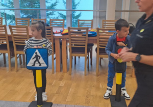 Dzieci trzymają znaki drogowe i sygnalizatory świetlne pod okiem pani ze straży miejskiej.