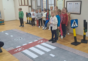 Dzieci stoją przy przejściu dla pieszych, a pani ze straży miejskiej tłumaczy im zasady bezpiecznego przechodzenia przez jezdnię.