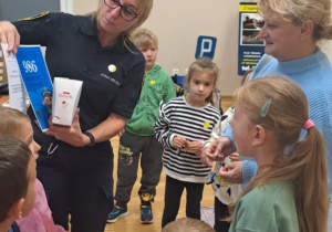 Pani ze straży miejskiej pokazuje dzieciom plakat numerem alarmowym, a w ręku trzyma upominek od dzieci.