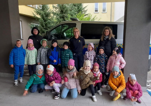 Dzieci pozują do zdjęcia grupowego wraz ze swoimi paniami oraz funkcjonariuszką straży miejskiej na tle radiowozu.