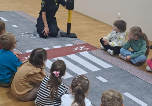 Pani ze Straży Miejskiej pokazuje dzieciom sygnalizator świetlny i uczy zasad przechodzenia przez ulicę.