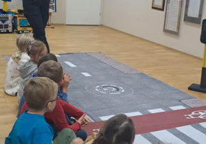 Pani ze straży miejskiej pokazuje dzieciom znaki drogowe podczas zajęć edukacyjnych.