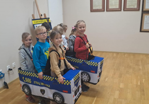 Dzieci biorą udział w zabawie edukacyjnej, udając radiowóz straży miejskiej.