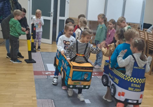 Dzieci bawią się w ruch drogowy, poruszając w materiałowych pojazdach i ucząc się zasad bezpiecznego przechodzenia przez jezdnię.