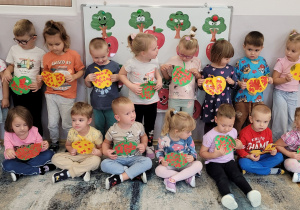 Dzieci z grupy Słoneczka siedzą i stoją na tle tablicy i prezentują swoje prace.