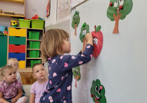 Dziewczynki i chłopcy siedzą na dywanie, a dziewczynka przyczepia na tablicę wydrukowane jabłko.
