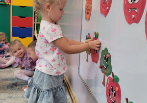 Dziewczynki i chłopcy siedzą na dywanie, a dziewczynka przyczepia na tablicę wydrukowane jabłko.