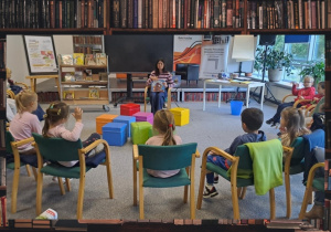 Dzieci z grupy Motylki siedzą na krzesełkach, przed nimi siedzi pani i czyta książkę.