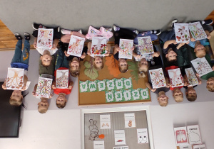 kilkoro dzieci stoi a kilkoro siedzi przy tablicy z dekoracją z okazji dnia drzewa i prezentują wykonane przez siebie prace plastyczne przedstawiające jesienne drzewo.