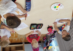 dzieci siedzące przy stoliku kolorują pień i gałęzie drzewa brązową kredką.