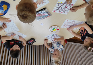 dzieci siedzące przy stoliku kolorują kredką pień i gałęzie drzewa, a patyczkami higienicznymi umoczonymi w kolorowej farbie stemplują kolorowe liście na drzewie.