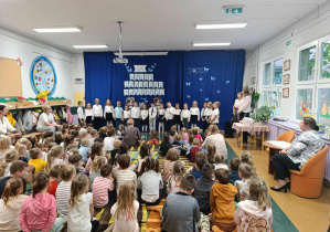 Dzieci ubrane na galowo stoją na tle dekoracji, przed nimi siedzą pozostałe dzieci oraz panie.