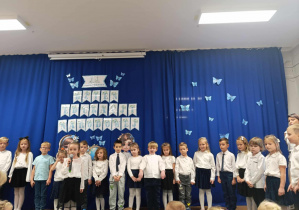 Dzieci stoją na tle dekoracji, jedna dziewczynka trzyma w ręku mikrofon.