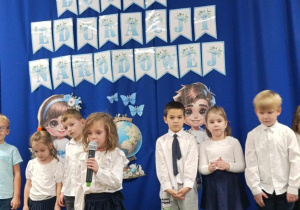 Dziewczynka trzyma mikrofon a reszta dzieci stoi na tle dekoracji.