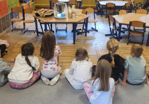 Dzieci siedzą na dywanie, słuchają jakie składniki potrzebne są do przygotowania chleba.