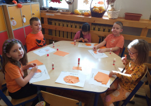 Zdjęcie przedstawia dzieci wykonujące pracę plastyczną - wyklejanie konturu dyni kawałkami pomarańczowego papieru.