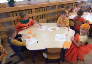 Zdjęcie przedstawia dzieci wykonujące pracę plastyczną - wyklejanie konturu dyni kawałkami pomarańczowego papieru.