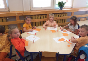 Zdjęcie przedstawia dzieci wykonujące pracę plastyczną - wyklejanie konturu dyni kawałkami pomarańczowego papieru.