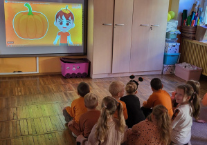 Dzieci siedzą na dywanie, oglądają film edukacyjny na tablicy multimedialnej pt. ,,Kazio i Dzień Dyni”.