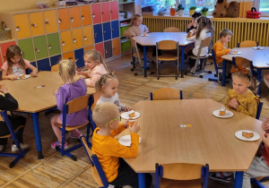 Dzieci siedzą przy stolikach, częstują się ,,pomarańczowym” ciastem.
