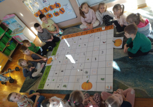 Na zdjęciu widzimy dzieci siedzące na dywanie oraz mate do kodowania na której rozłożone są różne poszczególne elementy dyni nos, oczy, usta. Dzieci mają przed sobą sylwetki dyni oraz roboty do kodowania.