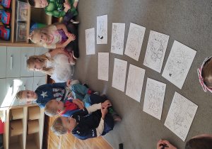 Dzieci siedzą na dywanie w kole, po środku którego rozłożone są kolorowanki z bajkowymi postaciami.