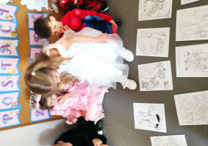 Dziewczynka przygląda się rozłożonym na dywanie bajkowym ilustracjom, w tle dzieci siedzące na dywanie