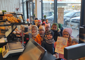 Przedszkolaki ustawione przy ladzie sklepowej pozują do zdjęcia, jedna dziewczynka trzyma w dłoniach podziękowania dla ekspedientki.