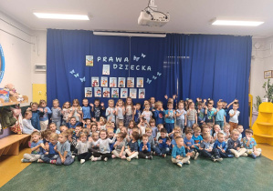 Przedszkolaki w grup najmłodszych pozują do zdjęcia na tle dekoracji z informacjami o prawach dziecka.