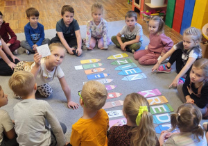 Dzieci siedzą na dywanie, dziewczynka nazywa co jest na rysunki i dopasowuje do kredki w odpowiednim kolorze.