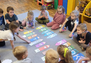 Dzieci siedzą na dywanie, dziewczynka nazywa co jest na rysunki i dopasowuje do kredki w odpowiednim kolorze.