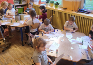Dzieci siedzą przy stolikach, wycinają kredki.