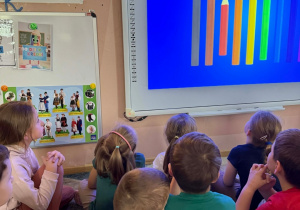 Dzieci oglądają na tablicy multimedialnej film edukacyjny dotyczący kredek i kolorów.