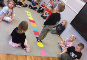 Zdjęcie przedstawia dzieci i dziecko spacerujące po dywanie utworzonym z figur geometrycznych.